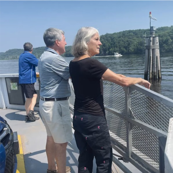 The image shows a ferry deck with two individuals: a woman in a black shirt and pants and a man in a light blue shirt and white shorts, both leaning on the metal railing. The background features a navigational buoy and green trees along the water's edge, with a sunny atmosphere and clear visibility.