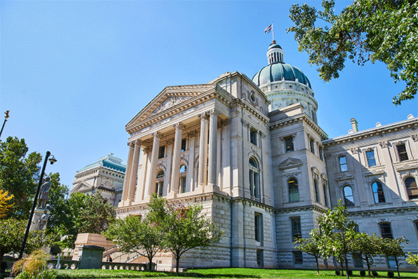 Outside image of Indiana State University’s Normal Hall, a stately white stone building, three stories high, with tall columns, wide steps, and a dome at the top. Blue sky is visible above, and the building is surrounded by green grass and several green trees.