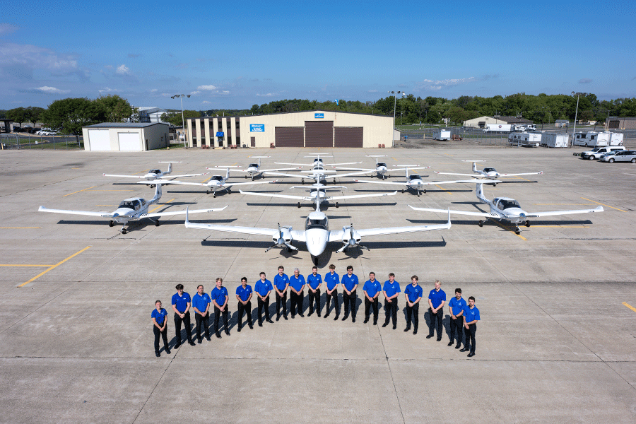 A group of people in matching blue polo shirts and black trousers stands in a curved formation on a large concrete tarmac. Behind them, a fleet of small white aircraft is neatly arranged. A beige hangar with brown doors and a blue sign stands in the background under a blue sky, surrounded by trees, buildings, and parked vehicles.  