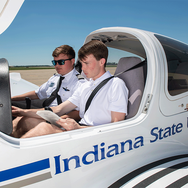 Two young white men with dark brown hair are seated inside an aircraft with an open canopy. One, wearing a white polo shirt, holds a checklist, while the other, dressed in a pilot's uniform and sunglasses, observes. Both are wearing seatbelts. The side of the aircraft displays “Indiana State” in blue. The background features a clear blue sky and a tarmac.