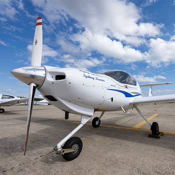 A white propeller aircraft with blue letters reading “Flying Sycamores Sydney Kremer” on its body sits on a tarmac under a bright blue sky with scattered clouds. Another white aircraft is partially  visible on the left  side.
