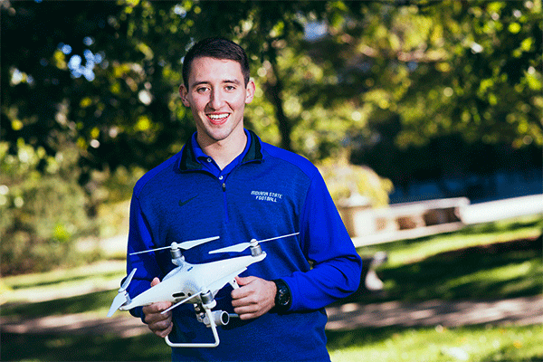 A smiling white man with short dark hair stands outdoors with greenery visible behind him. He wears a blue zip-up jacket that says “Indiana State University” and holds in both hands a white drone with three propellers on it.  