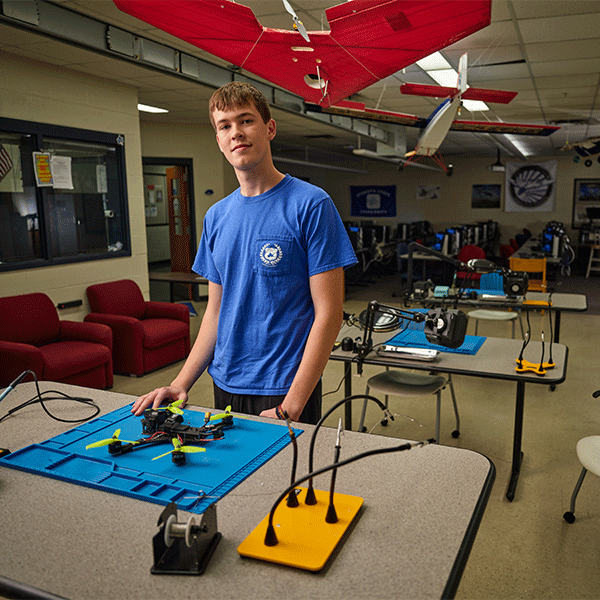 A young white man with short light brown hair in a blue t-shirt stands in a classroom, resting his hand on a small drone on a blue mat. He is smiling slightly and exudes confidence. The room contains various tools and workstations. Several model aircraft with red wings hang from the ceiling. The background shows computers, workstations, and red lounge chairs.
