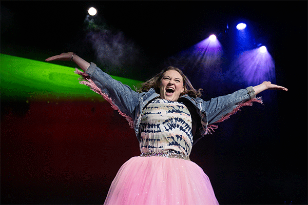 A young white woman with shoulder-length dark brown hair stands on a stage with outstretched arms, wearing a blue jean jacket, a colorful patterned shirt, and a fluffy pink skirt. She smiles excitedly under colorful stage lights.