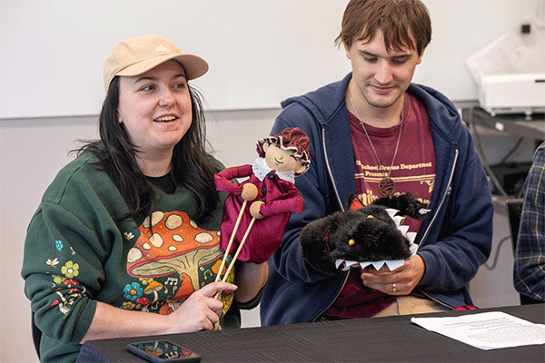 A young white woman with dark brown hair, wearing a beige cap and a green sweater with a mushroom drawing, sits at a black table and smiles while holding a puppet outfitted in red. Next to her, a young white man with blond hair smiles slightly. He wears a red T-shirt and a navy-blue hoodie, and he holds a black monster puppet with yellow eyes and sharp teeth.