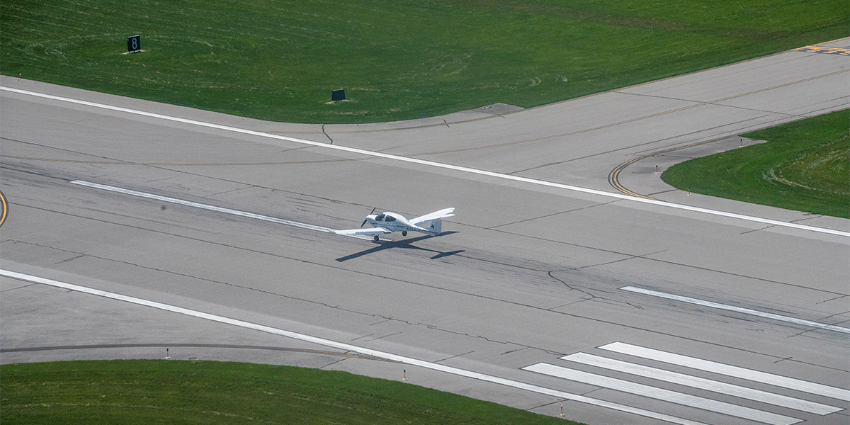 An aerial view of a small white aircraft taxiing on a wide runway. The gray tarmac has visible tire marks, white guiding lines, and a large runway number. Green grass borders the runway, from all sides. In the distance, intersecting taxiways and signage mark the airport's layout.