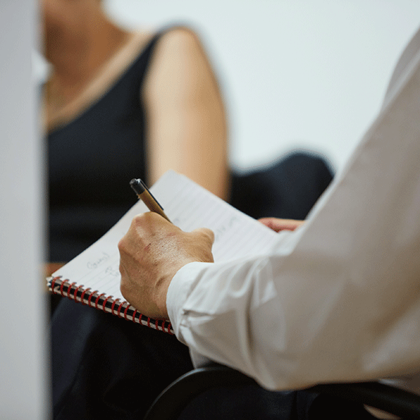 A person in a white long-sleeved shirt writes in a spiral-bound notebook with a pen. Another person, partially visible and wearing dark clothing, sits across from them.
