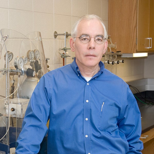 Dr. Arthur Halpern stands in a laboratory setting. He has white hair and round glasses, and he wears a blue button-up shirt with a pen in the shirt pocket. Behind him are seen various scientific instruments, including gauges and glassware, mounted on the wall and countertop. The background also features wooden cabinets and tiled walls.