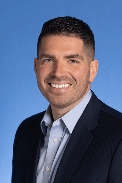 Kent Games A smiling man with short, black, neatly styled hair and a goatee is wearing a dark suit jacket over a light blue dress shirt. He stands against a solid blue background. The person's hair is short and neatly styled.