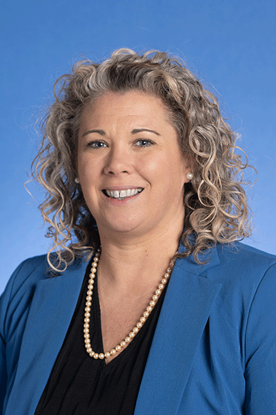 Dr. Lindsey Eberman A smiling woman with curly, gray-blonde hair wears a blue blazer, black top, and pearl necklace, She stands against a solid blue background.