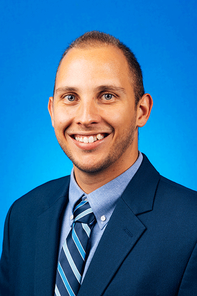 Dr. Matthew Rivera A smiling man in a dark blue suit jacket, light blue-striped dress shirt, and blue-striped tie, stands against a solid bright blue background.