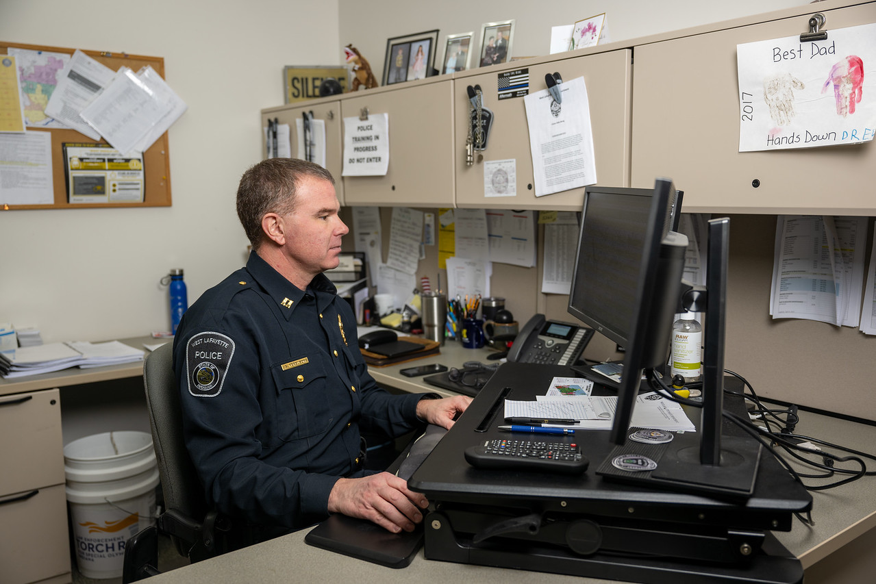 A middle-aged white male police officer in a dark navy uniform sits at a desk in an office, working on a computer with papers, notes, and family drawings pinned to the cabinets around him.