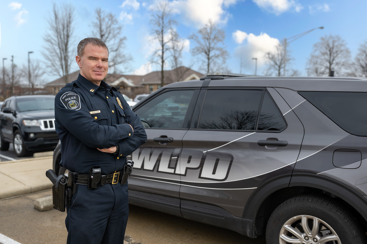 A middle-aged white male police officer in a dark navy uniform stands with arms crossed in front of a gray Lafayette Police vehicle marked “WLPD.”
