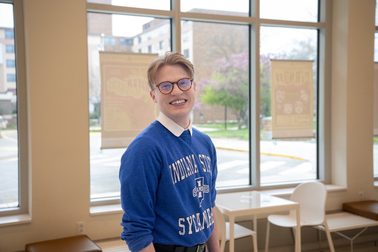 A young white man with short blond hair and glasses smiles while standing indoors near large windows. He is wearing a blue “Indiana State Sycamores” sweatshirt over a white collared shirt. Outside the windows, trees and a building are visible.