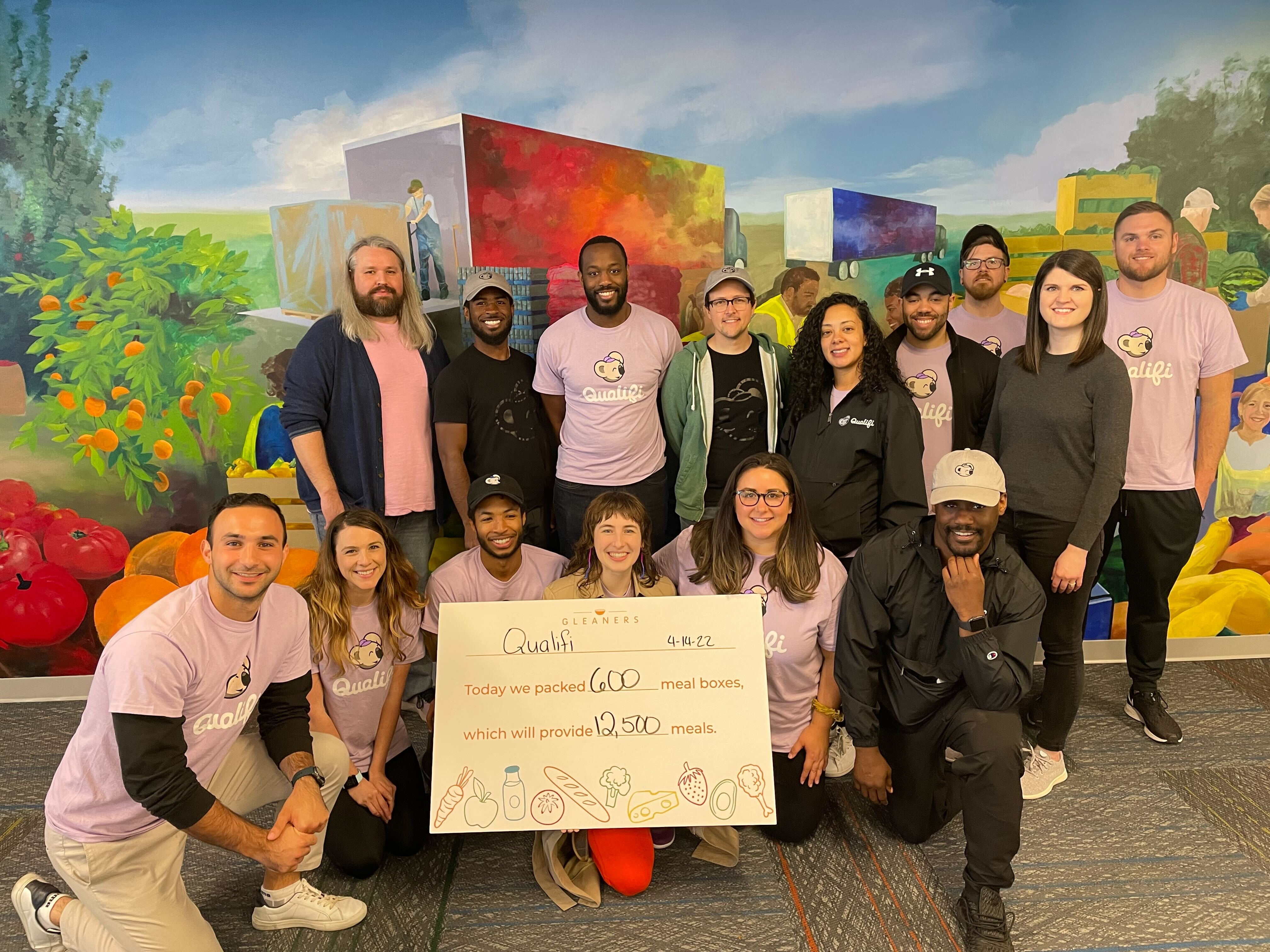 A diverse group of men and women pose together indoors in front of a colorful mural. Many wear light purple “Qualifi” shirts, while others are dressed in casual clothing. The group holds a large sign that reads: “Qualifi — Today we packed 600 meal boxes, which will provide 12,500 meals” with the date 4-14-22.