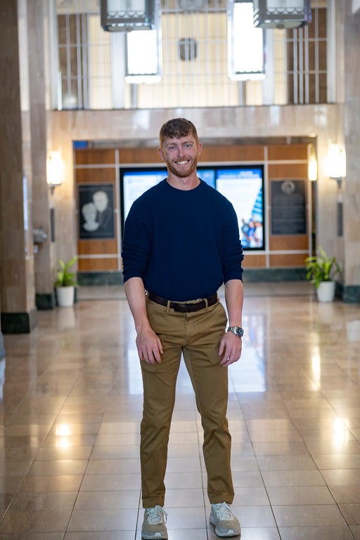 A young white man with short curly hair and a beard smiles while standing in a bright hallway with polished floors. He is wearing a navy long-sleeve shirt, khaki pants, and white sneakers. Behind him are digital screens, framed portraits, and potted plants.