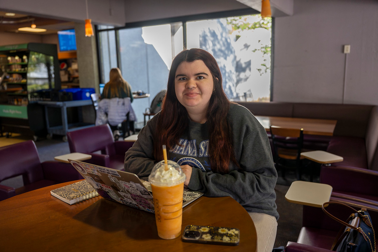 A young white woman with long dark hair sits at a table in a café, wearing a gray Indiana State University sweatshirt. In front of her are a laptop, a notebook, a phone, and a large orange Starbucks drink topped with whipped cream.