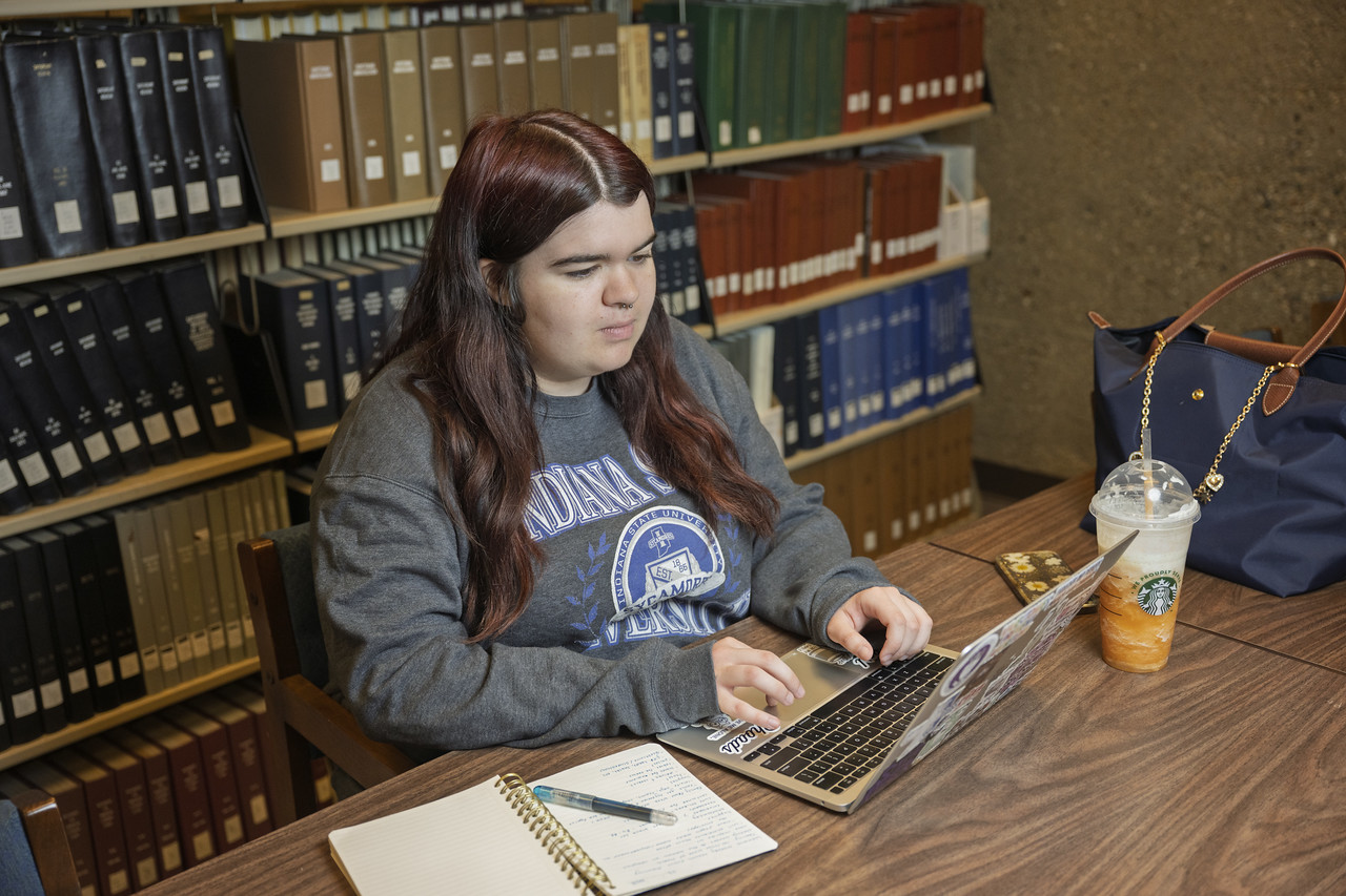 A young white woman with long dark hair sits at a wooden table in a library, working on a laptop. She is wearing a gray Indiana State University sweatshirt, with a notebook, pen, and a Starbucks drink on the table beside her.