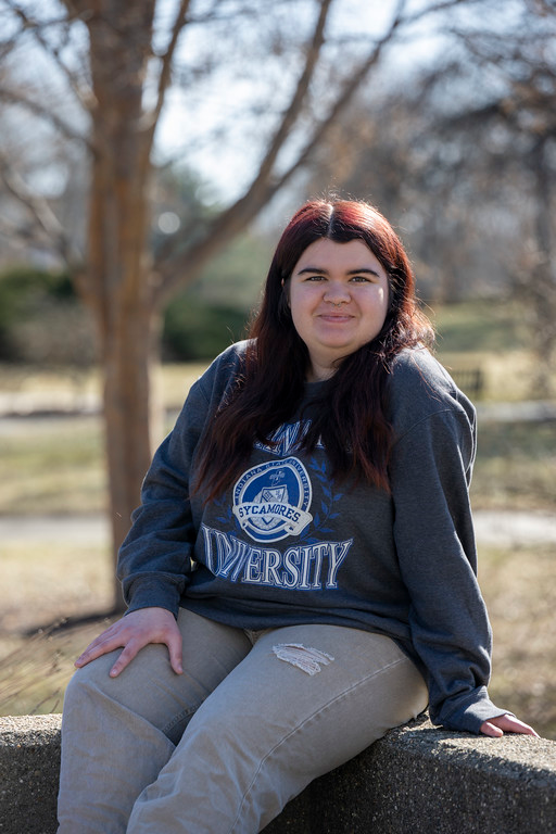 A young white woman with long dark hair sits outdoors on a low concrete ledge, wearing a gray Indiana State University sweatshirt and light-colored pants, with trees and grass in the background.