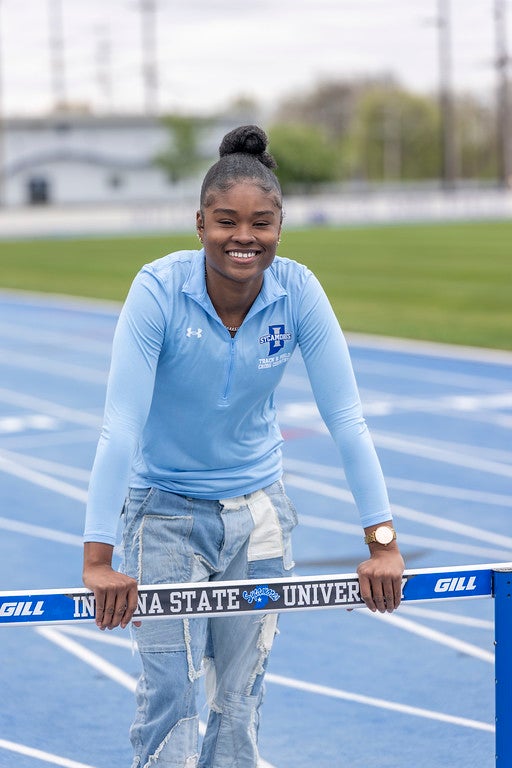 A young Black woman in a light blue long-sleeve shirt with an Indiana State Sycamores logo and patchwork jeans smiles while leaning on a hurdle that reads “Indiana State University.” A blue running track and grassy field are in the background.