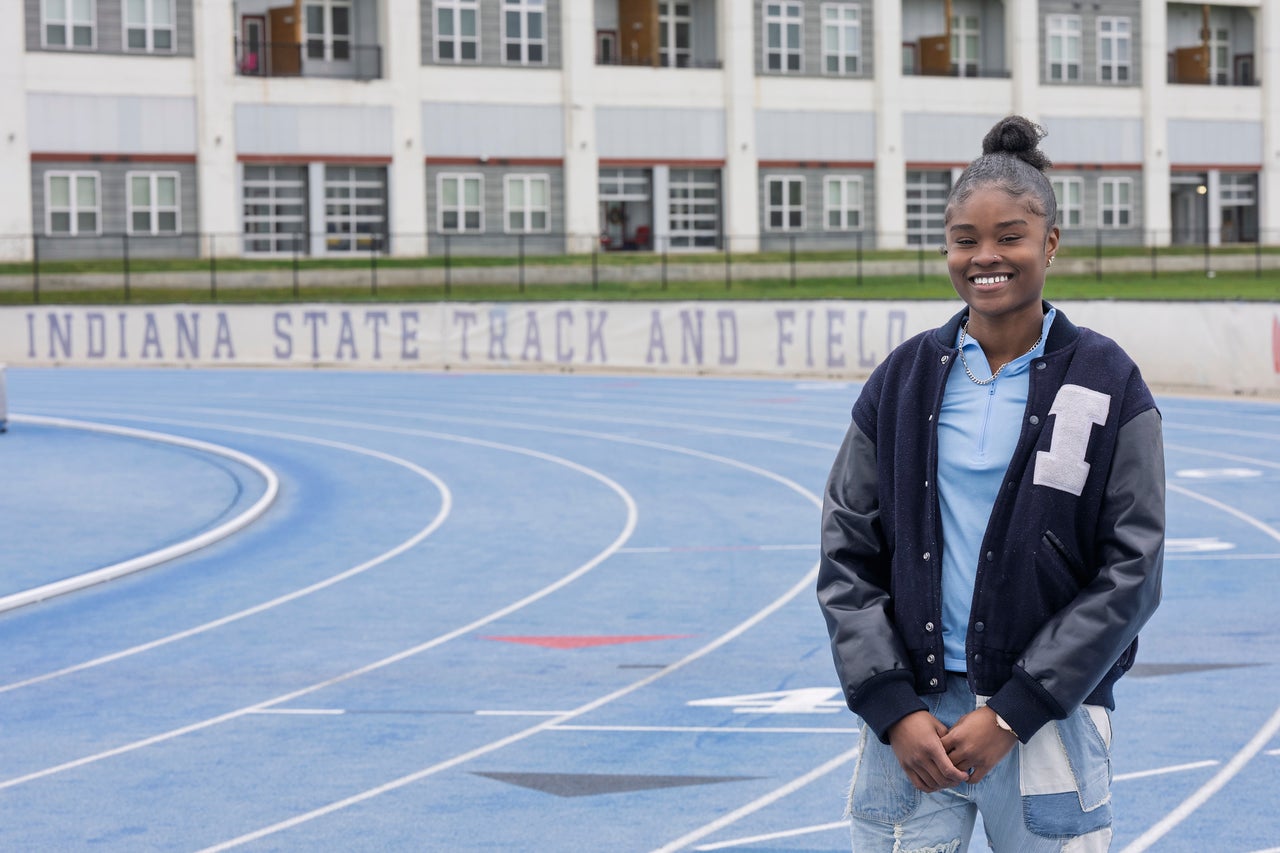 A young Black woman smiles while standing on a blue running track. She wears a navy letterman jacket, a light blue shirt, and jeans. Behind her, a banner reads “Indiana State University Track and Field,” with a building in the background.