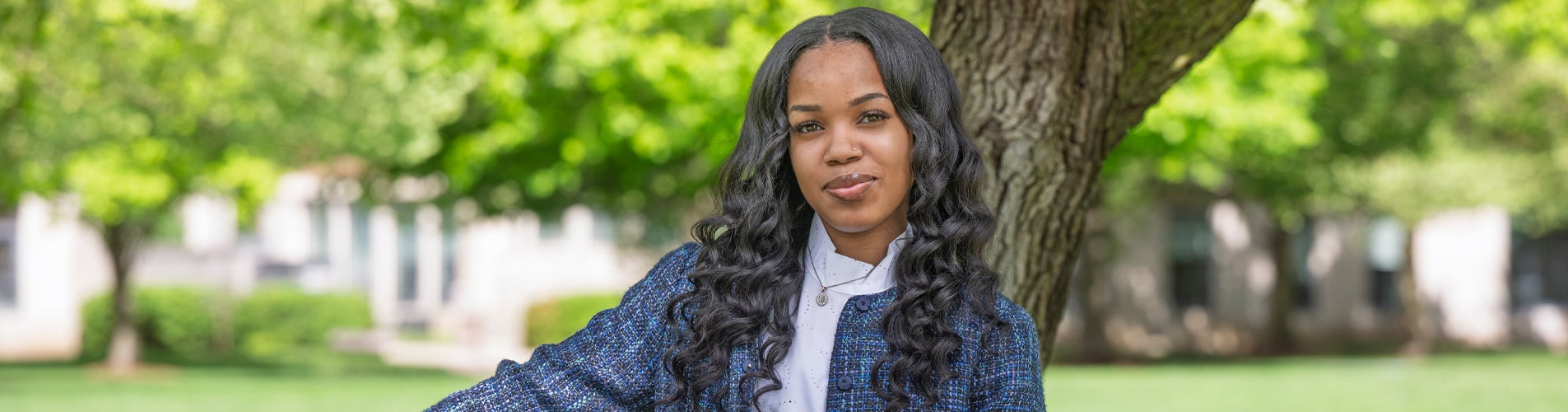 A young Black female with long, wavy hair is standing outdoors. She is wearing a white shirt and a blue textured jacket. The background features a tree behind her, and a blurred grass and foliage..