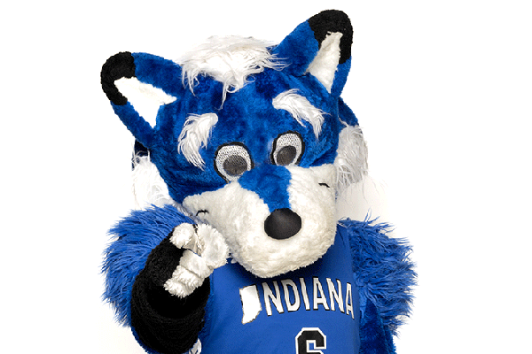 A blue and white mascot, a woodland creature named Sycamore Sam, points outward toward the viewer with a slight tilt of the head. The mascot is furry with big ears. It wears an Indiana State University jersey.