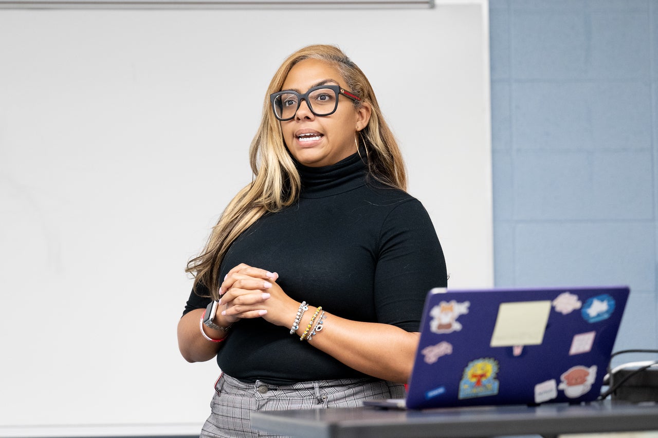 A Black woman with long blonde hair and large black glasses stands in front of a whiteboard, speaking with her hands clasped. She wears a black turtleneck and plaid pants, with a laptop covered in colorful stickers on the desk beside her.