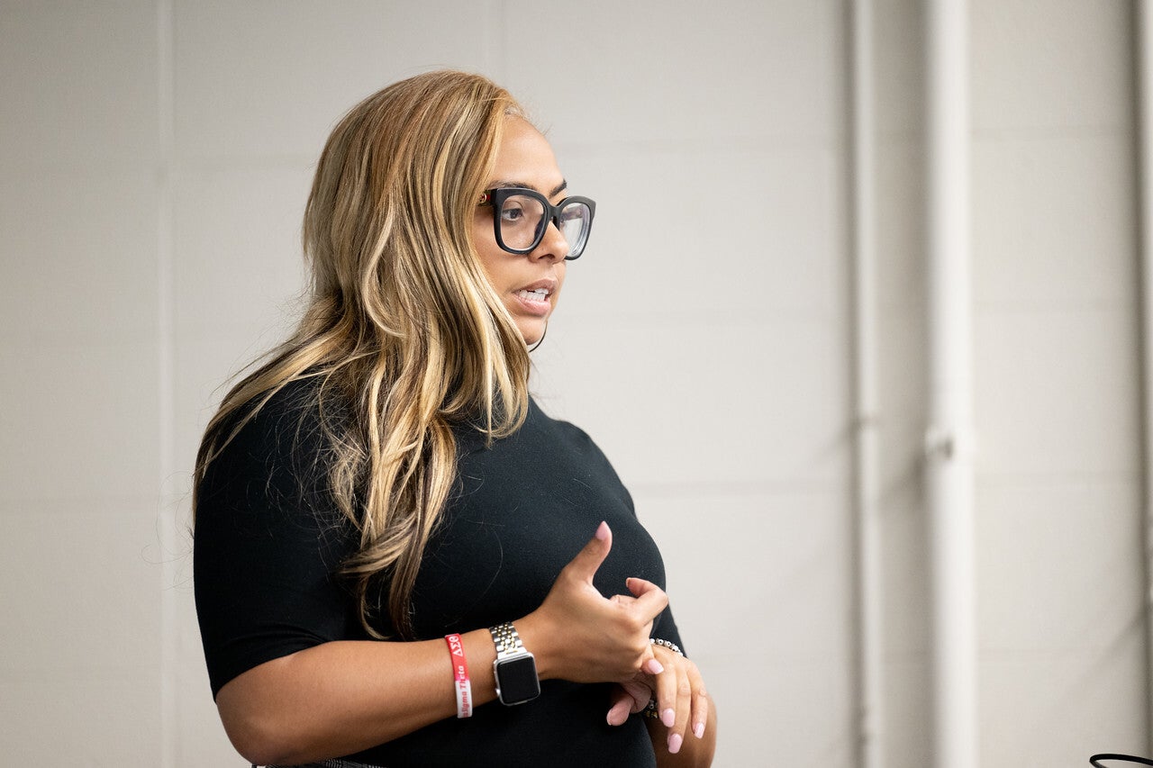 A Black woman with long blonde hair and large black glasses speaks while gesturing with her hands. She wears a black top, a watch, and a red wristband, standing against a light-colored wall.