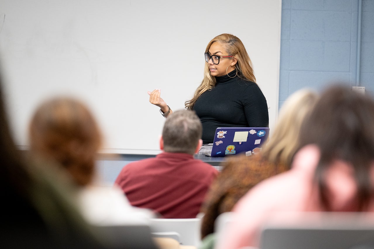 A Black woman with long blonde hair and large black glasses speaks to a classroom of students. She wears a black turtleneck and hoop earrings, standing beside a laptop covered in colorful stickers.