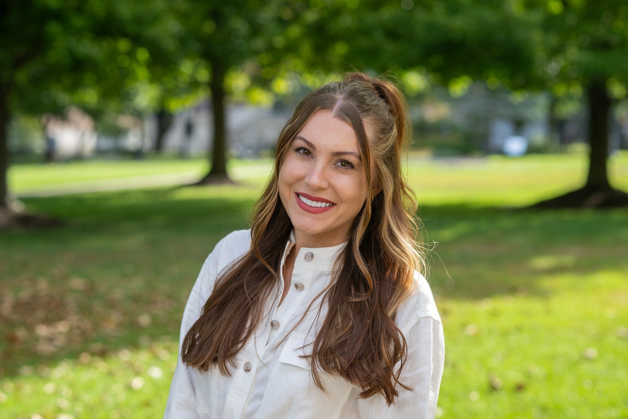 White woman with long brown wavy hair stands outdoors smiling in a sunlit area, wearing a white button-up shirt, with trees with green leaves and well-kept green grass visible in the background.