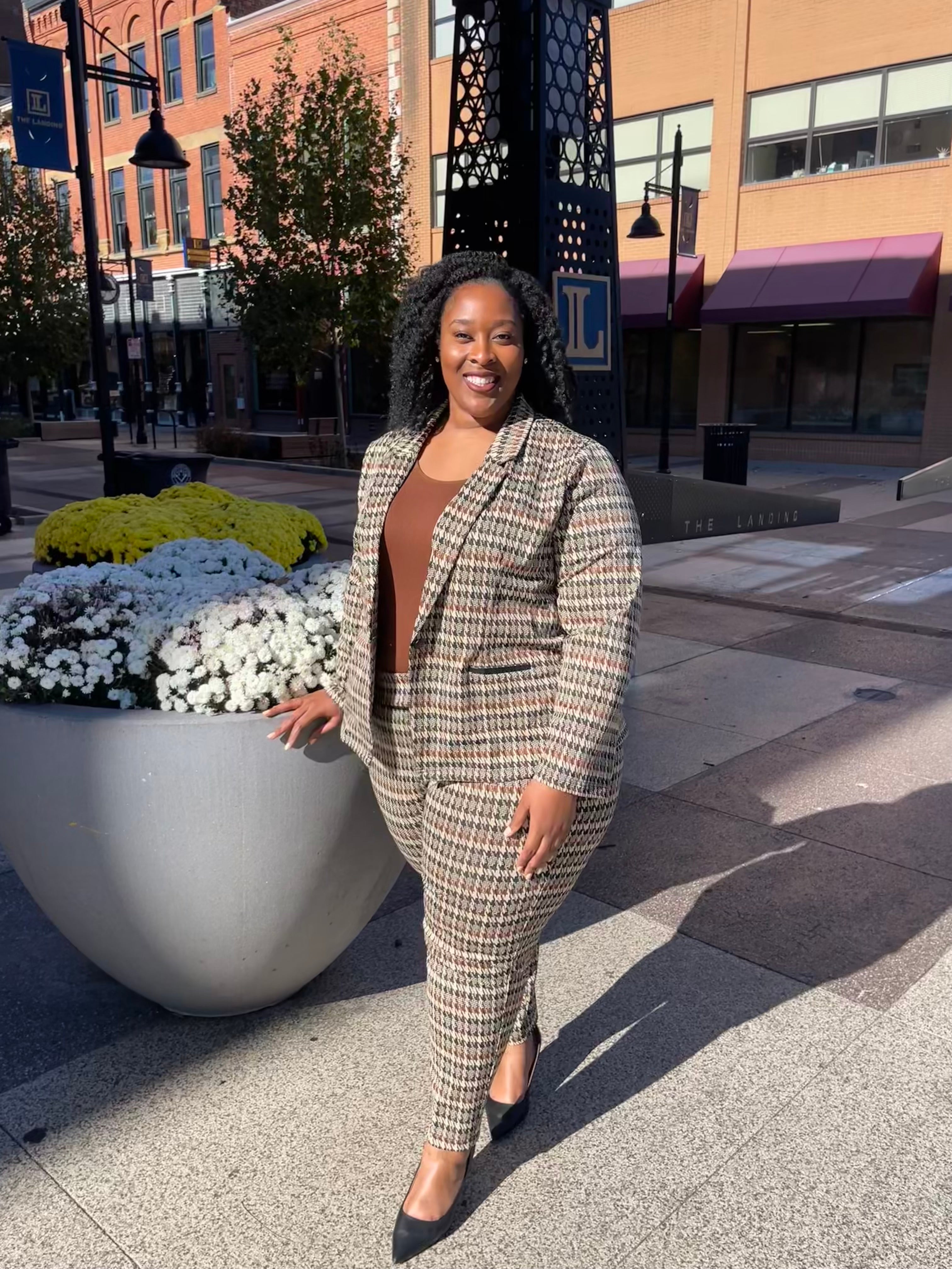 A smiling Black woman stands outdoors on a sunny day, wearing a brown top with a matching plaid blazer and pants. She poses beside a large planter of white and yellow flowers, with brick buildings and street lamps in the background.