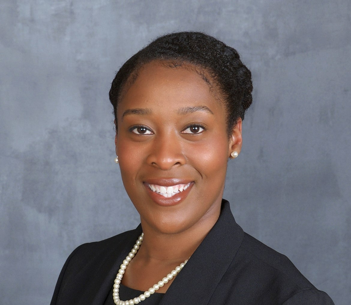 A smiling Black woman wearing a black blazer, pearl necklace, and pearl earrings poses against a gray background.