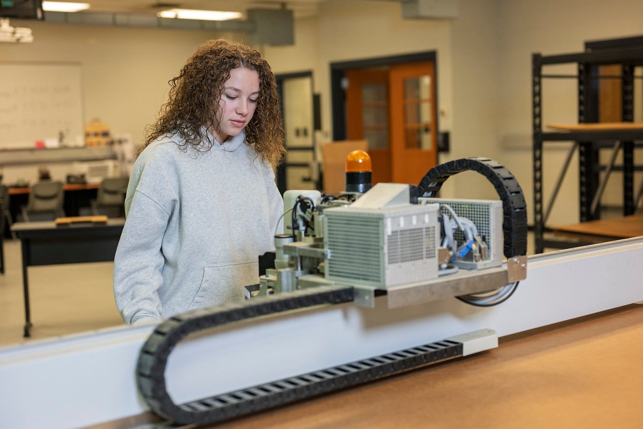 A female with curly brown hair wearing a light gray hoodie stands in a workshop or lab, observing or operating a machine with a long track and an orange warning light. The background includes shelves, tables, chairs, and various equipment typical of an industrial setting.