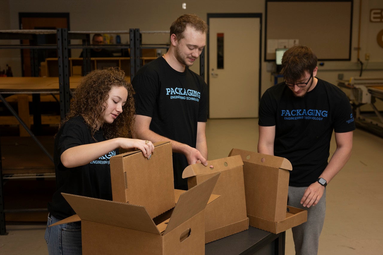 Three individuals wearing black t-shirts labeled 'PACKAGING ENGINEERING TECHNOLOGY' are assembling cardboard boxes in a room with industrial shelving and equipment. One person is folding a box, another is holding a partially assembled box, and the third is looking down at another box. The setting resembles a training environment focused on packaging engineering.