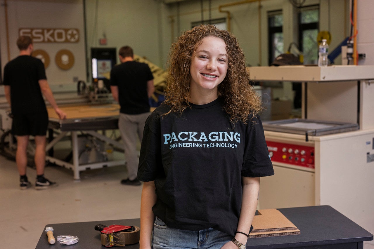 A female with curly brown hair is smiling and standing in a workshop, wearing a black t-shirt that reads 'PACKAGING ENGINEERING TECHNOLOGY.' In the background, three other individuals are engaged with machines and equipment. The workshop contains tools, machinery, and workbenches, creating an industrious, technical atmosphere.