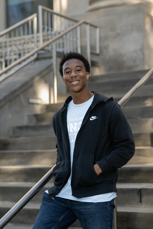 A young Black man wearing a black Nike zip-up hoodie over a light blue T-shirt and blue jeans stands smiling on an outdoor staircase in front of a stone building.