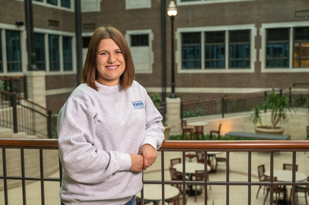 A young White woman with straight brown hair, wearing a gray “Honors College” sweatshirt, leans against a railing and smiles inside a brick academic building with tables, chairs, and potted plants visible in the background.