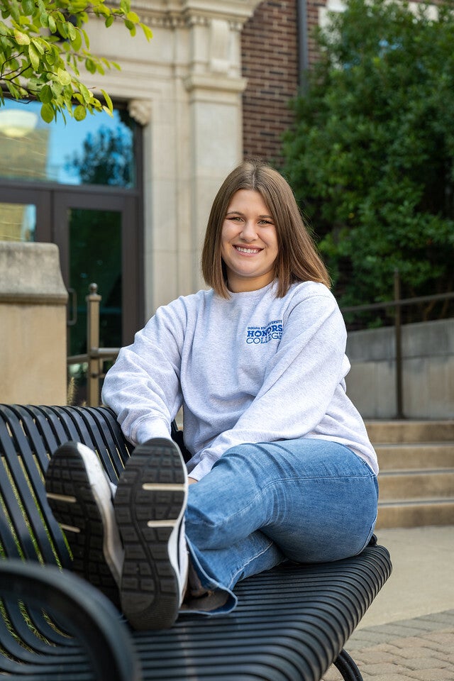 A young White woman with straight brown hair, wearing a gray “Honors College” sweatshirt, blue jeans, and black-and-white sneakers, sits smiling on a black metal bench with her legs crossed and stretched out outside a building.