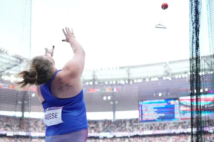 A female athlete in a blue outfit stands in a stadium filled with spectators, captured mid-action during a hammer throw. The hammer is airborne in the upper right corner. Her back is visible, showing a label with the name “REESE.” In the background, a large digital screen displays event information alongside flags from various countries.