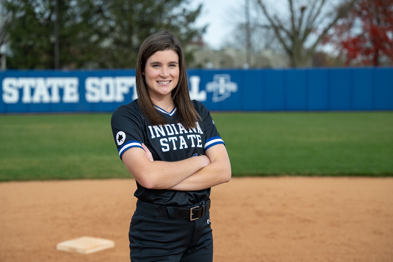 A young White woman with straight brown hair stands on a softball field with her arms crossed, wearing a black Indiana State University softball uniform with blue and white trim. The blue “Sycamores Softball” banner is visible in the background.