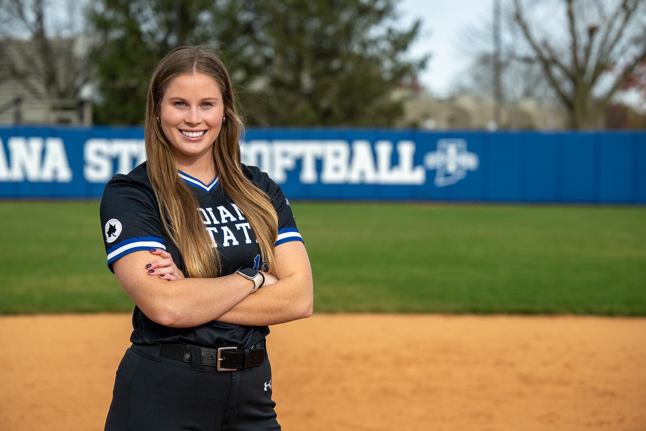 A young White woman with long straight blonde hair stands on a softball field with her arms crossed, wearing a black Indiana State University softball uniform with blue and white trim. The blue “Sycamores Softball” banner is visible in the background.