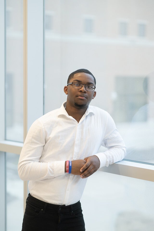 A Black man wearing glasses, a white collared shirt, and dark pants leans against a window ledge, looking at the camera with a calm expression.
