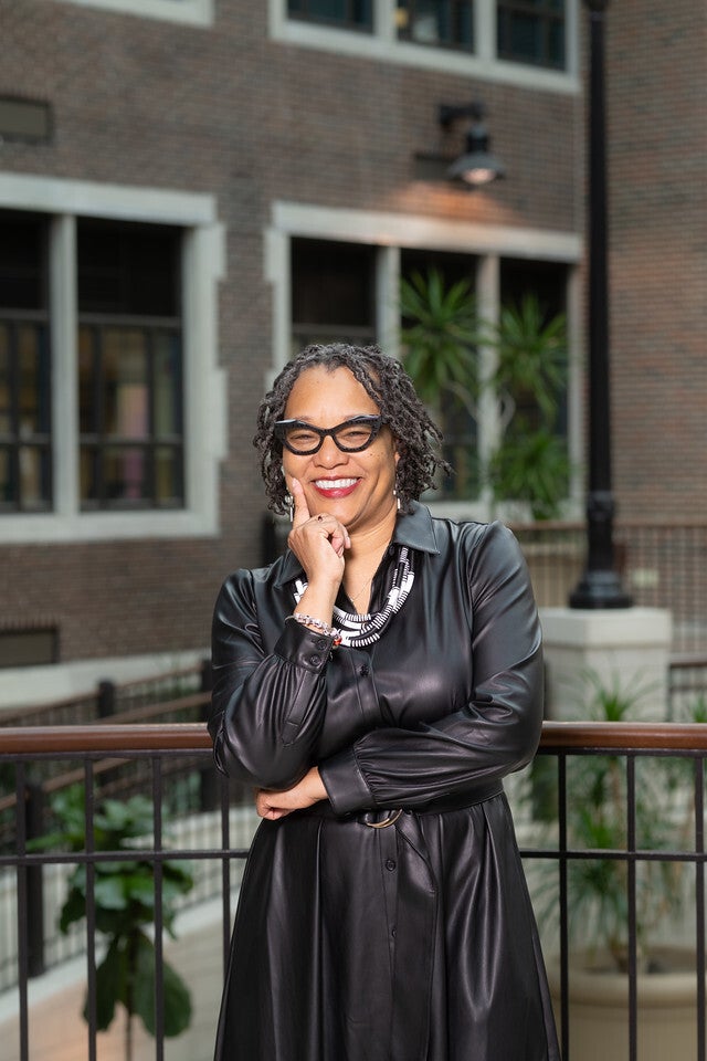 A smiling Black woman with short twists and black glasses stands indoors wearing a long black dress and a black-and-white beaded necklace. She leans on a railing with one hand resting on her chin. A brick building and green plants are visible in the background.