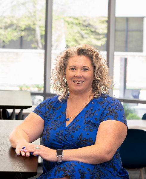 A smiling White woman with shoulder-length curly blonde hair sits at a table in a bright room with large windows. She wears a royal blue dress with a black floral pattern, a silver smartwatch, and a blue pendant necklace.