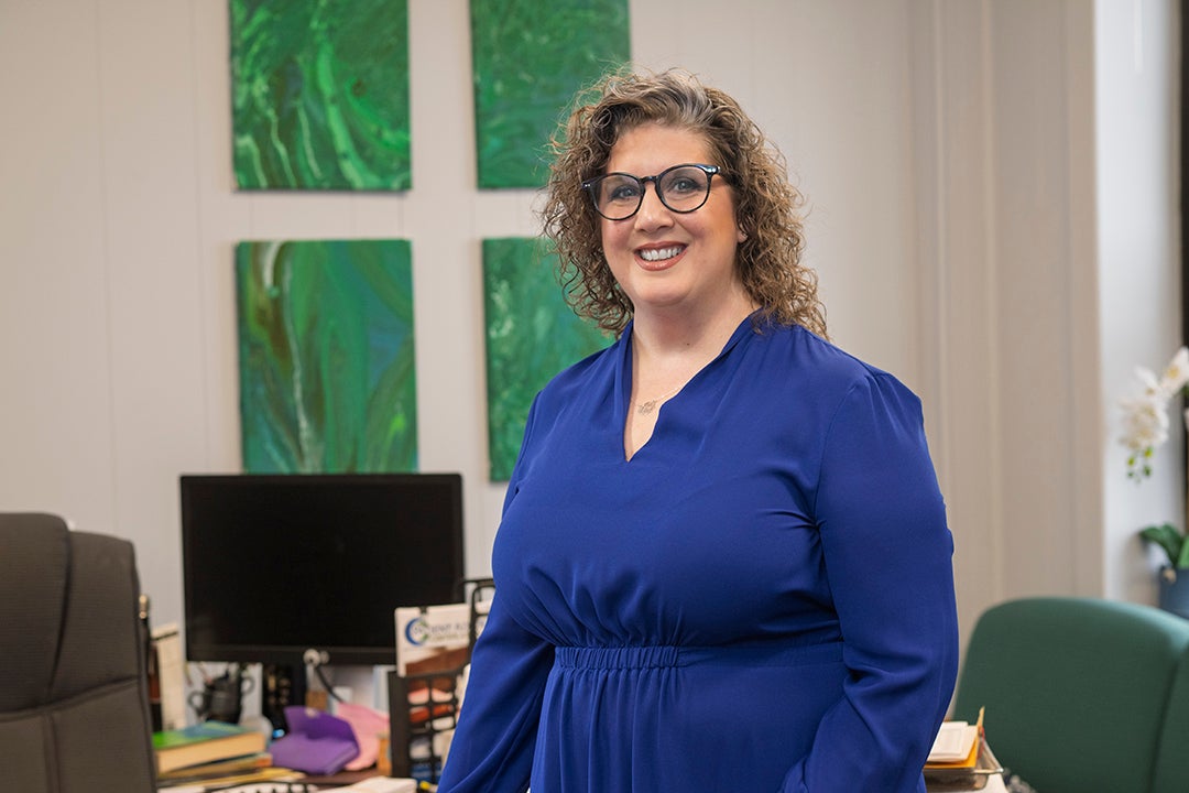A white woman with light brown curly hair and glasses, wearing a royal blue dress, stands in a professional office with a desk, computer, and green abstract art on the wall behind her