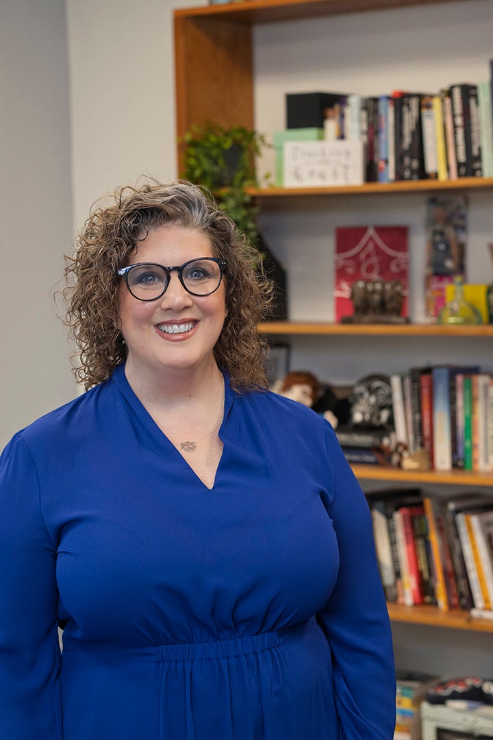 A white woman with curly light brown hair and glasses, wearing a royal blue blouse, stands in front of a bookshelf filled with books, decorative items, and a small green plant.