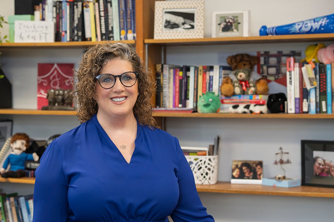 A white woman with curly light brown hair and glasses, wearing a royal blue blouse, stands in front of a bookshelf filled with books, framed photos, stuffed animals, and decorative items