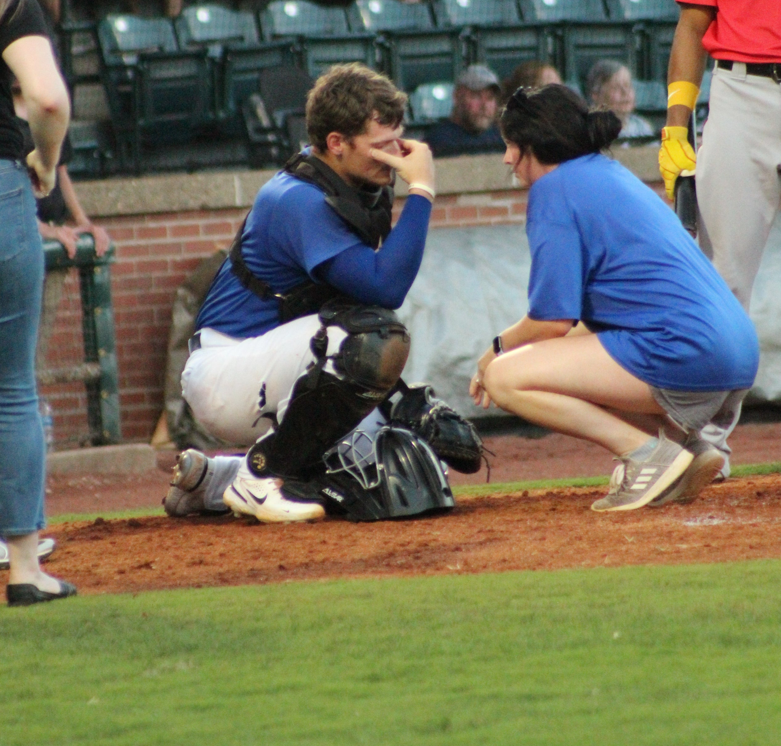 A White woman in a blue shirt and gray shorts kneels beside a White baseball catcher who crouches with his hand on his face while others look on nearby.