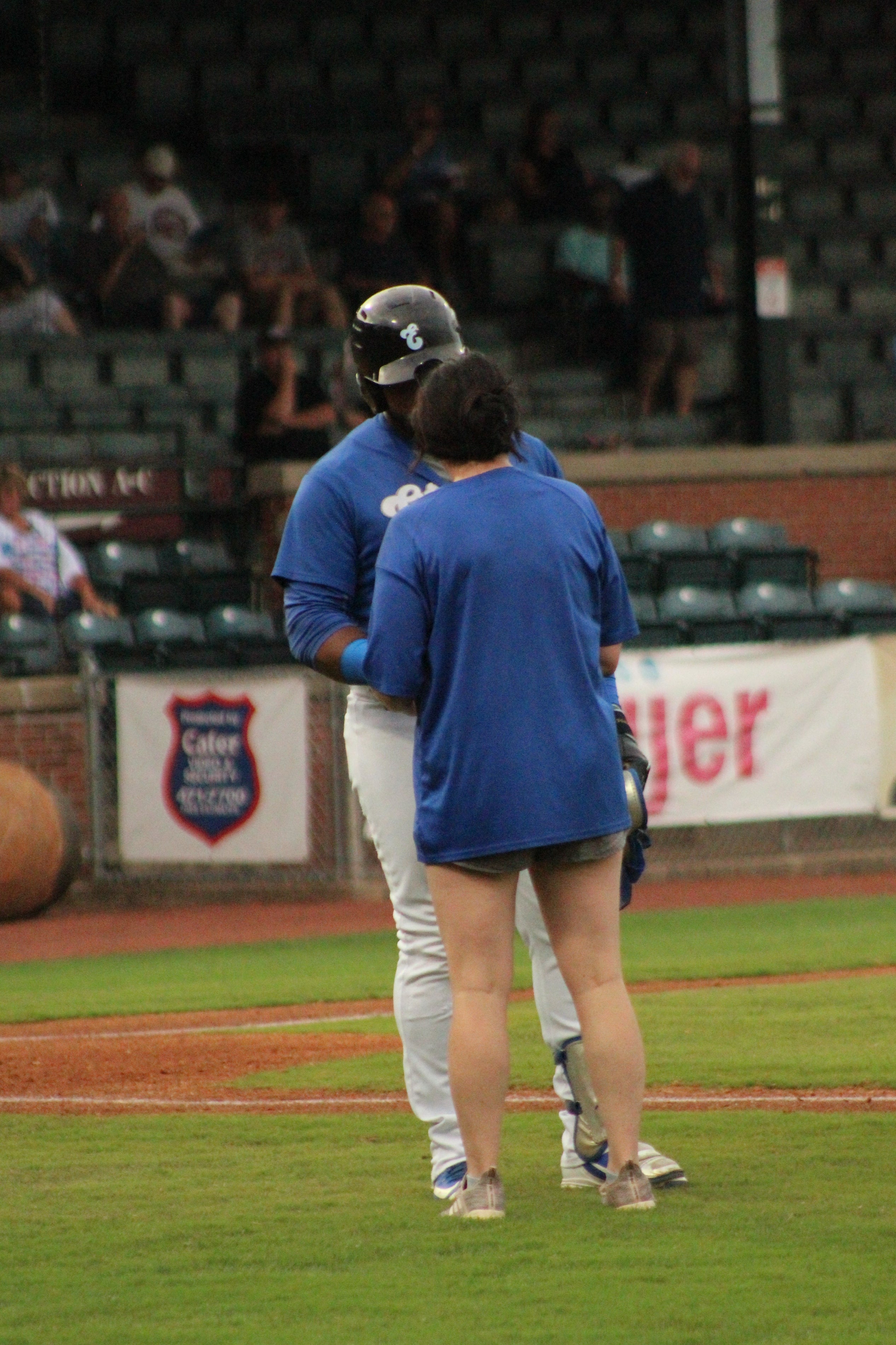 A White woman in a blue shirt and gray shorts stands on the field speaking with a Black baseball player in a blue uniform and helmet as they face each other near home plate.